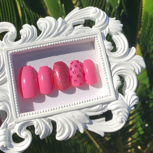 Pink press on nails with pink glitter on one nail and pink gems on another nail.